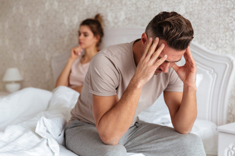 Sad man sitting on bed with his girlfriend on background at home